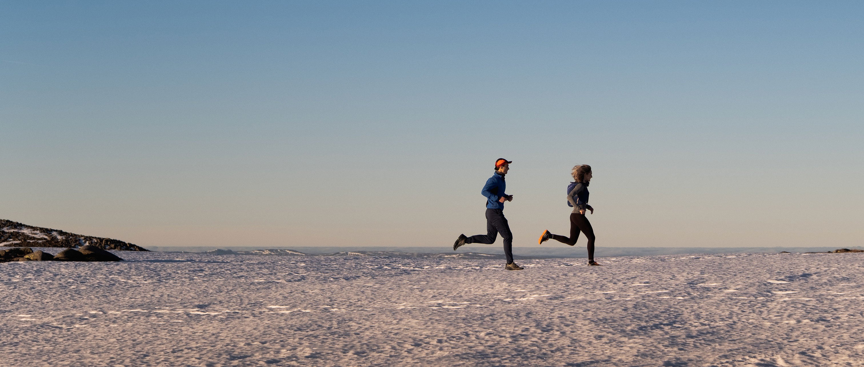 Two people running on a snowy landscape with a clear blue sky.