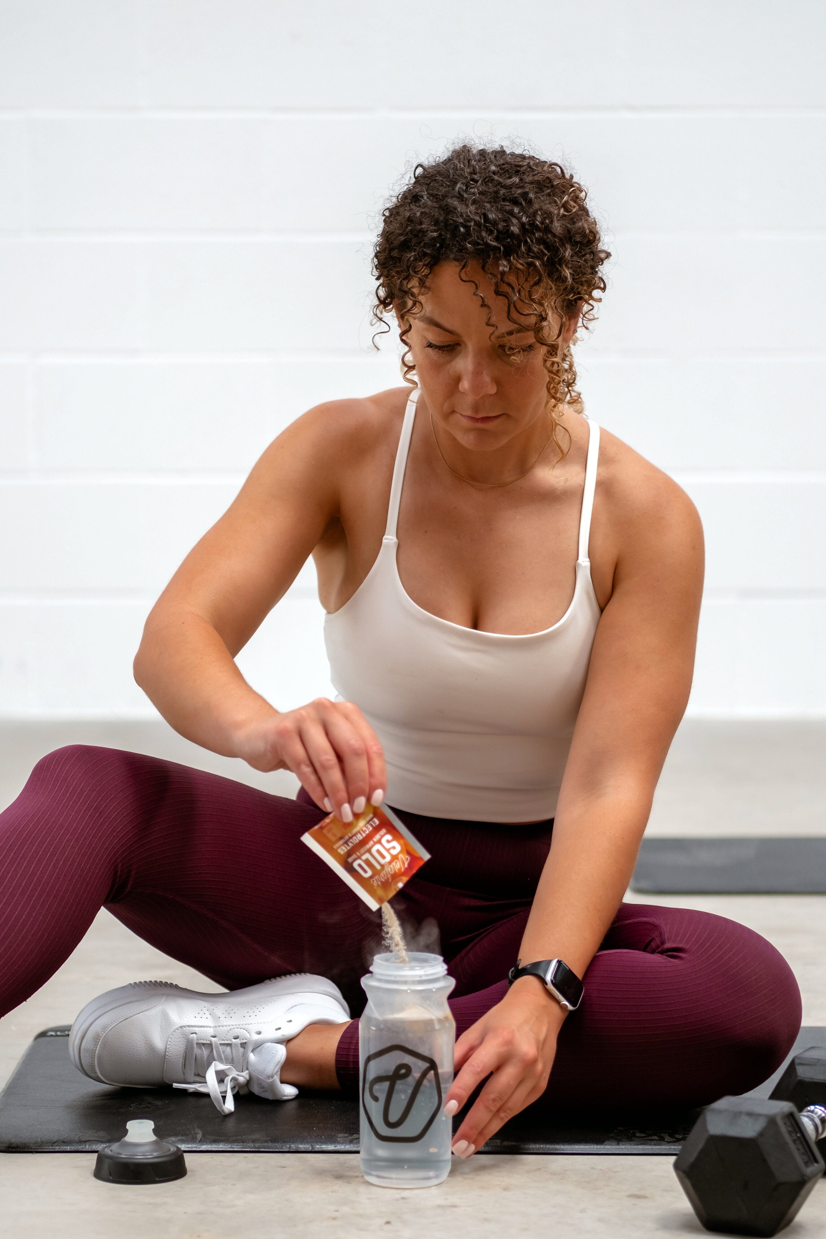 Woman sitting on the floor of a gym pouring Veloforte Electrolyte powder into a water bottle 