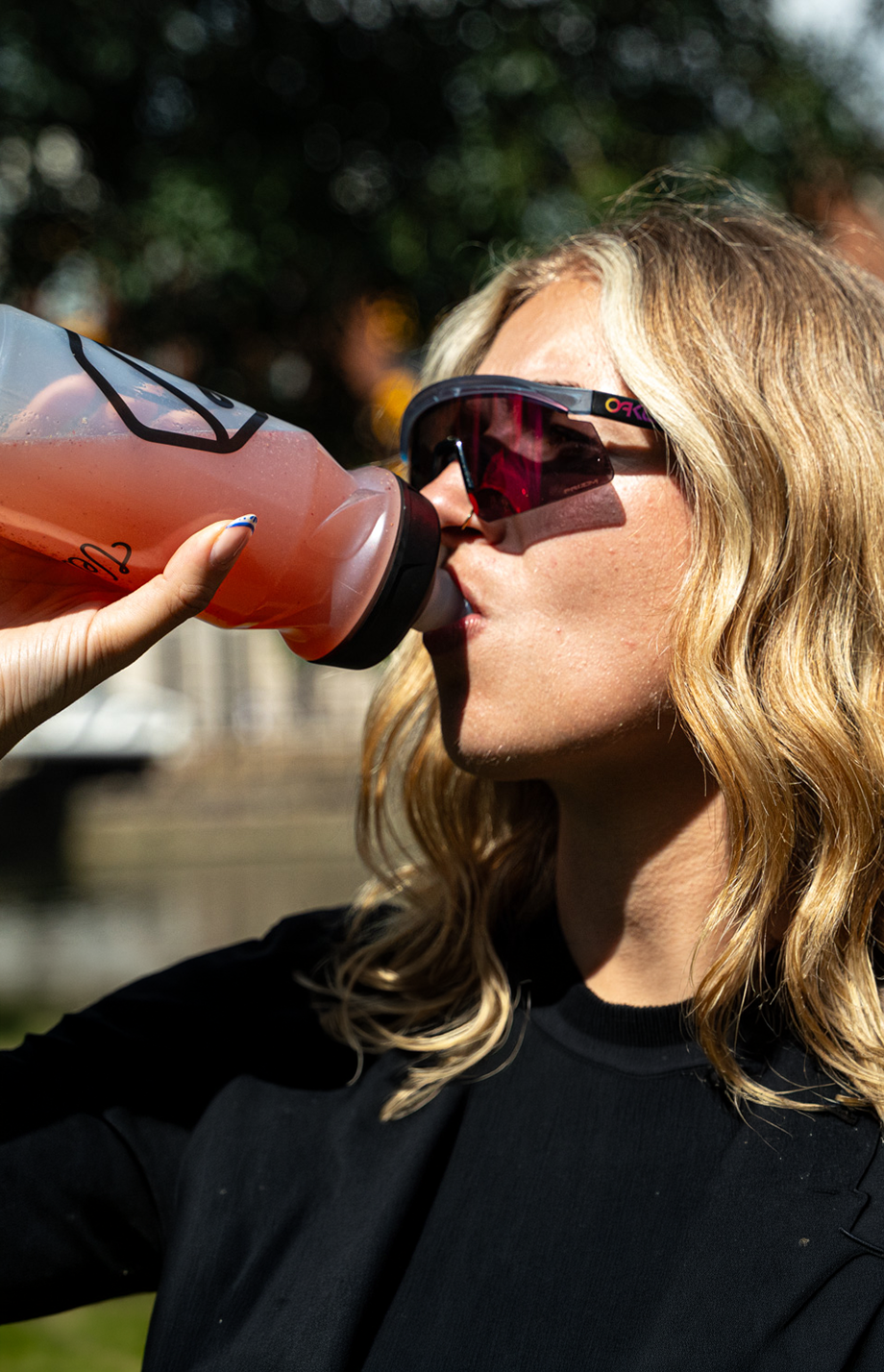 Person drinking from a pink bottle with sunglasses on, outdoors.