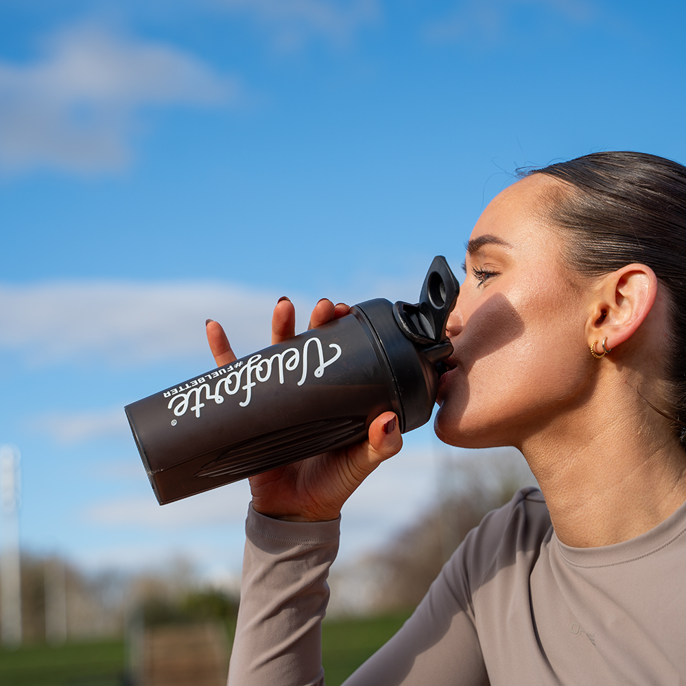 Person drinking from a black Veloforte protein shaker outdoors with a blue sky background
