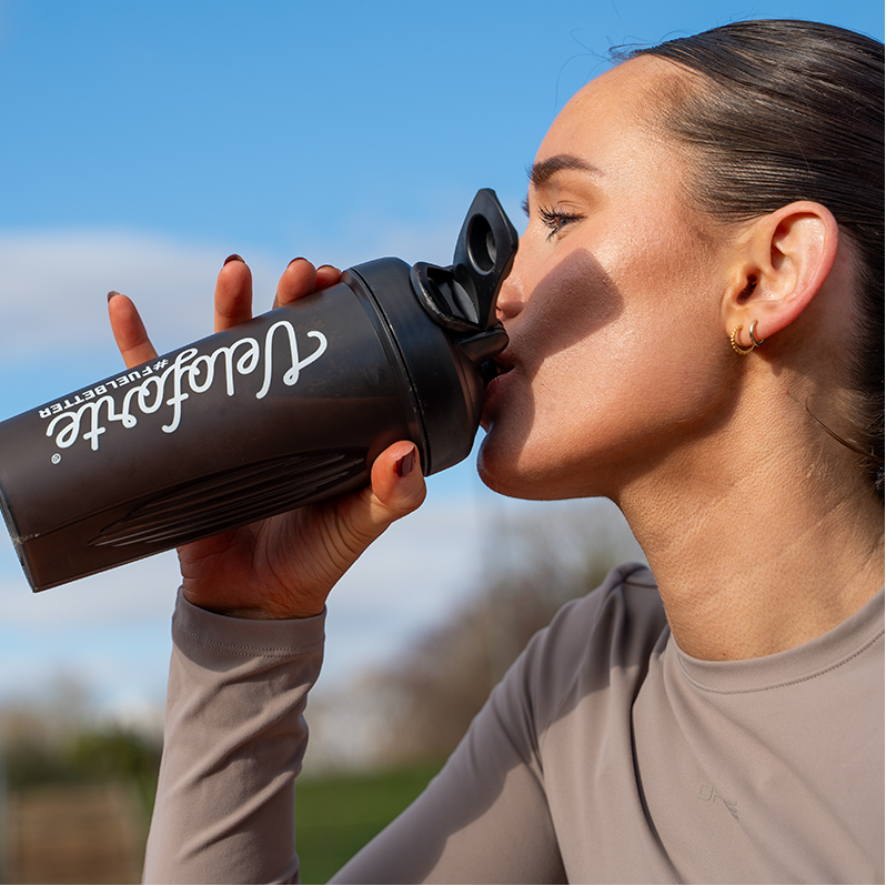 Person drinking from a black Veloforte protein shaker outdoors with a blue sky background