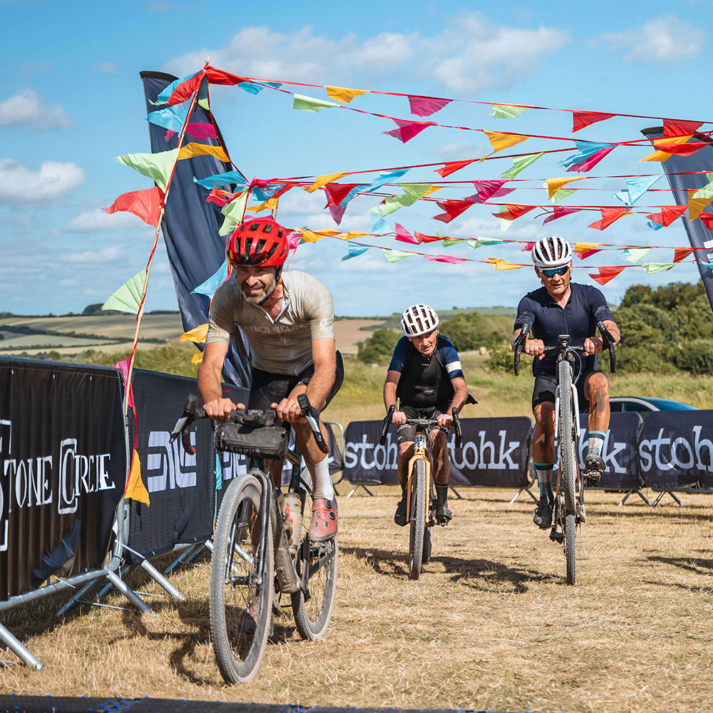 Three cyclists racing on a dirt track with colorful flags and banners in the background.