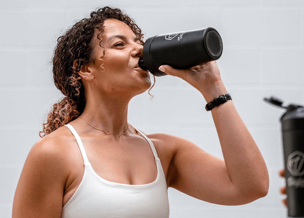 Woman in white tank top drinking from a black water bottle against a light gray background